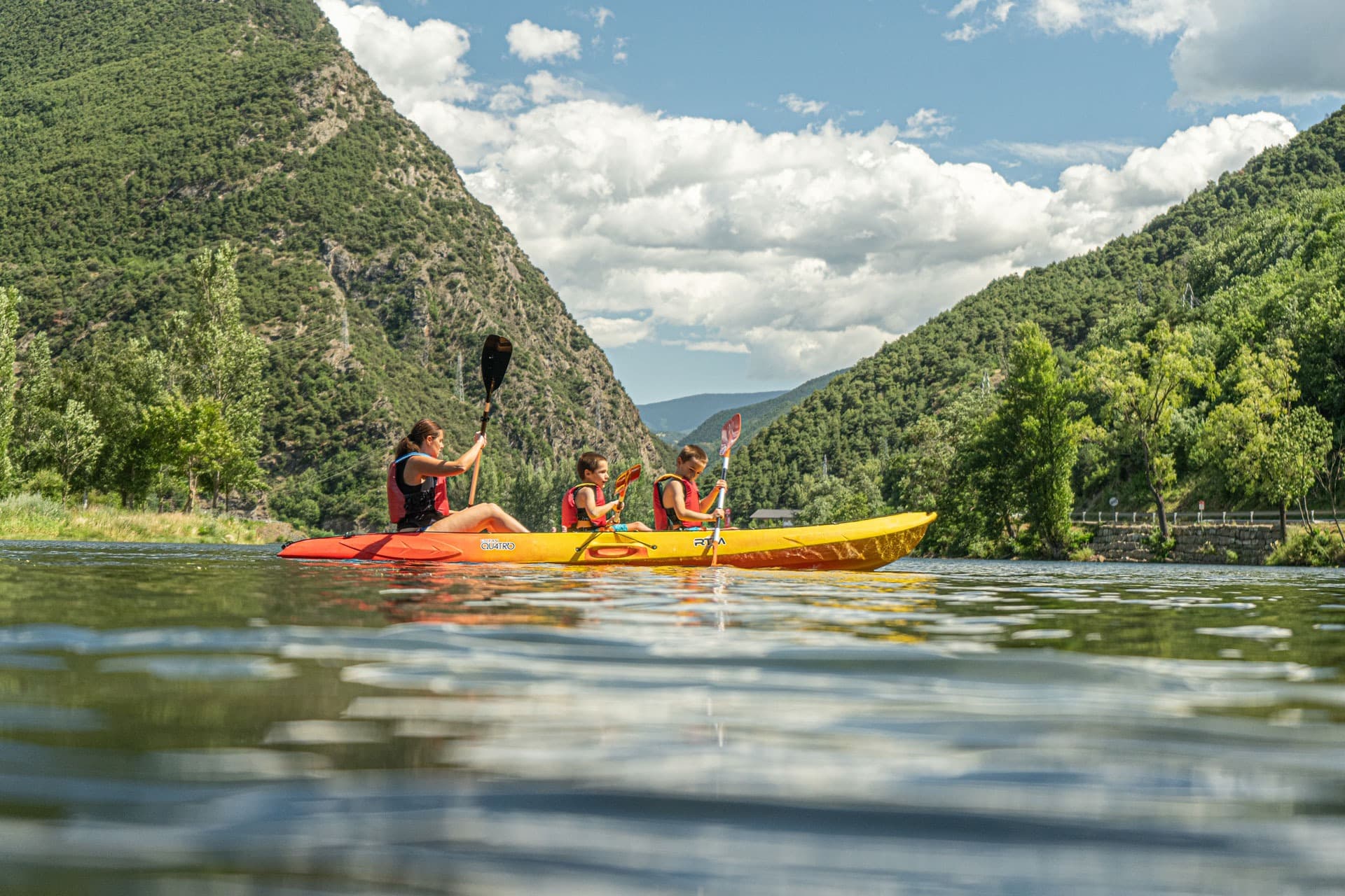 Several people paddling in single and double kayaks across the calm waters of La Torrassa Lake, surrounded by greenery.